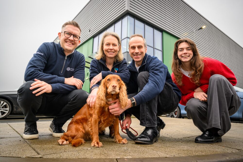 Four people crouching beside a golden-brown spaniel dog on a paved surface in front of a modern building with green-trimmed windows and grey corrugated metal cladding